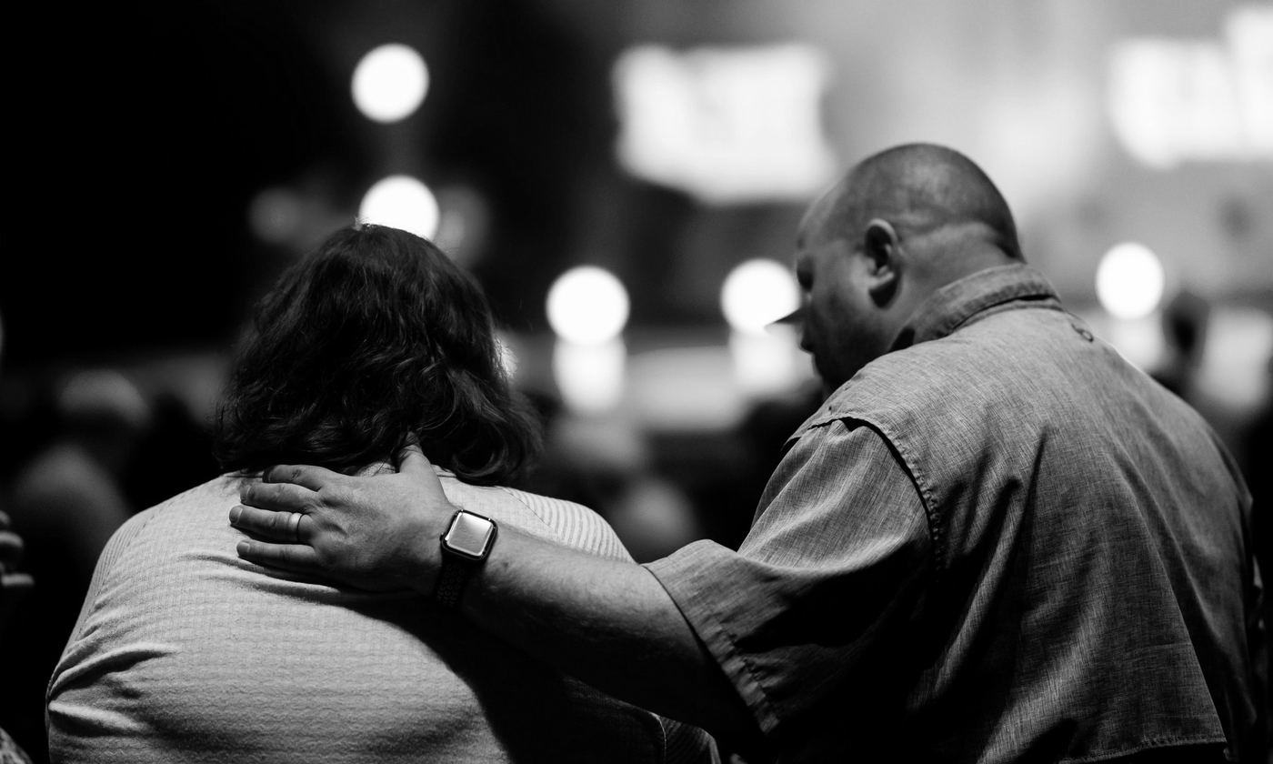 En una iglesia con luces de escenario al fondo, un hombre con la cabeza baja pone la mano en el hombro de la persona que está a su lado