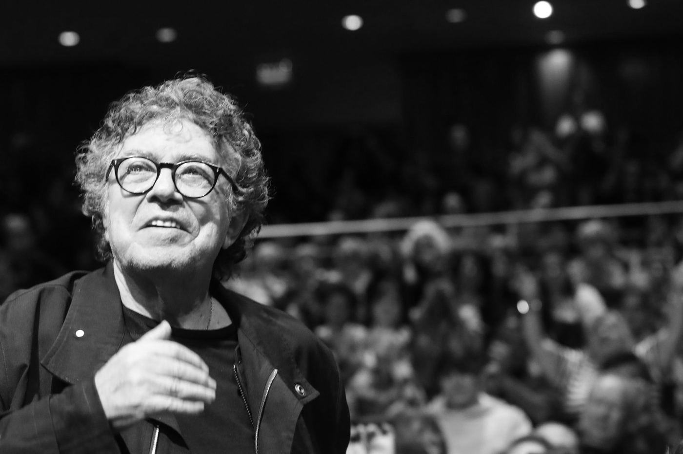 Piero, de pelo gris rizado y gafas, sonríe con la mano en el pecho frente a un auditorio lleno