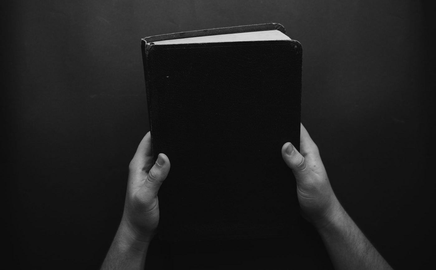 Dos manos sostienen en alto una Biblia negra y cerrada, sobre un fondo oscuro
